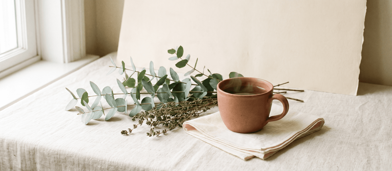 Eucalyptus and thyme sprigs beside a teacup with steam on cream linen